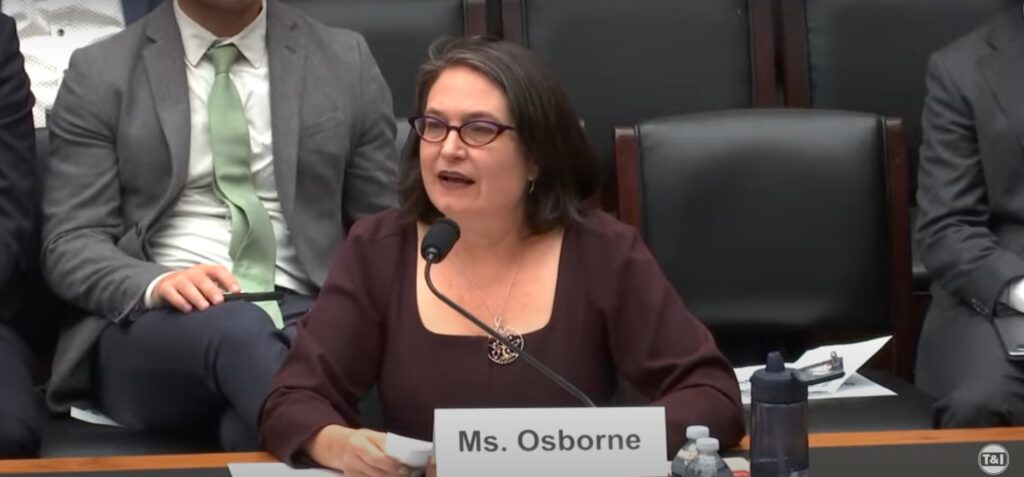 A woman with shoulder-length dark hair wearing glasses and a maroon top speaks into a microphone