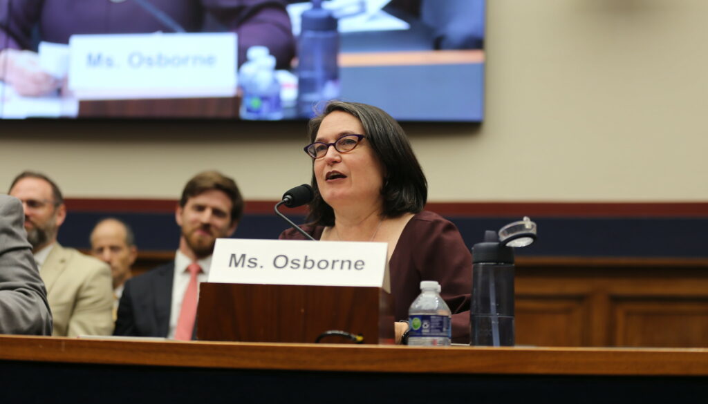 A woman with shoulder-length dark hair wearing glasses and a maroon top speaks into a microphone. Behind her are wooden benches and a yellow wall