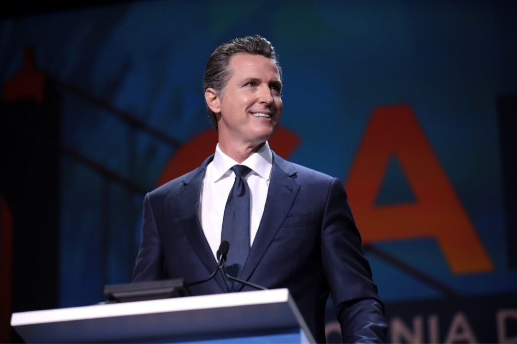 Governor Gavin Newsom wears a blue suit and tie, smiling from a podium