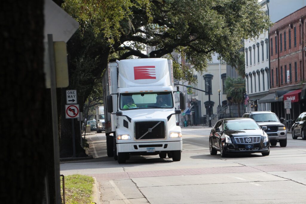 A large freight vehicle travels down a city street amid smaller vehicle traffic